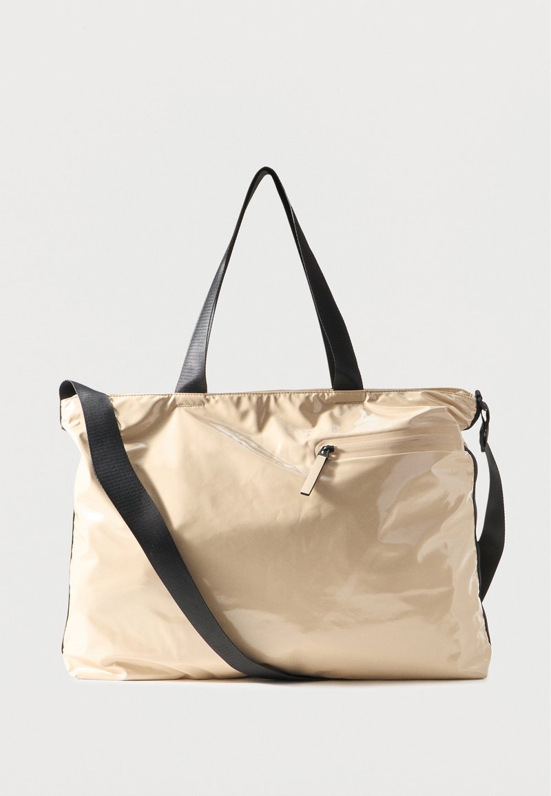 Μπεζ γυαλιστερή μεγάλη τσάντα ώμου με μαύρο ιμάντα ώμου και χερούλια, μπροστινή τσέπη με φερμουάρ, τοποθετημένη σε απλό ανοιχτόχρωμο φόντο.