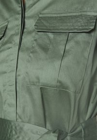 Tessuto verde con due tasche sul petto, dettagli di cucitura e una cintura annodata attorno alla vita di un indumento.