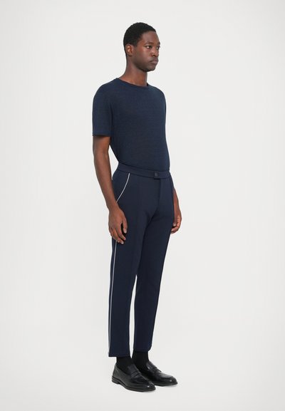 Marineblaues T-Shirt mit kurzen Ärmeln, kombiniert mit eng anliegenden marineblauen Hosen mit weißen Seitenstreifen. Das Modell trägt schwarze Mokassins und hat einen neutralen Gesichtsausdruck.