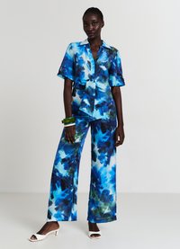 Completo due pezzi astratto nei toni del blu e del verde, composto da una blusa annodabile in vita e pantaloni a gamba larga. Tessuto liscio, maniche corte e sandali con tacco bianchi.