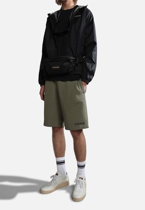 Chaqueta impermeable negra, pantalones cortos verde oliva, zapatillas blancas con suelas beige. Cintura con cordón y riñonera de marca en la cintura.