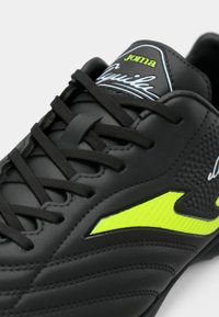 Μαύρο αθλητικό παπούτσι με υφή από συνθετικό υλικό, διαθέτοντας φωτεινές κίτρινες λεπτομέρειες και μαύρα κορδόνια. Περιλαμβάνει λογότυπο στη γλώσσα.