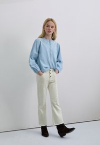 Camisa de mezclilla azul claro con cuello redondeado y cierre de botones, combinada con pantalones blancos de talle alto con botones frontales y dobladillos deshilachados.