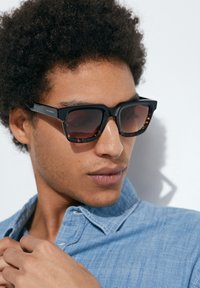 Ochelari de soare negri cu rame dreptunghiulare, având accente din material cu model țestoasă pe laturi. Purtați cu o cămașă din chambray albastră, cu textură netedă.