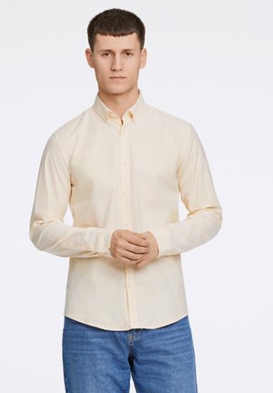 Camisa de botón de manga larga de color amarillo claro con collar, que presenta una textura suave y un dobladillo recto, combinada con unos vaqueros de mezclilla azules.