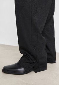 Black trousers featuring a subtle textured pattern with buttons at the ankle, paired with sleek black shoes. The fabric is smooth and matte.