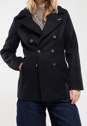 Kvinde iført en sort dobbeltradet uldfrakke over en grå striktrøje og mørkeblå bukser, stående med den ene hånd i lommen.