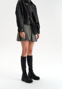 Czarne skórzane kozaki sięgające za kolano z grubą podeszwą. Modelka ma na sobie szaro teksturowaną spódnicę i czarną skórzaną kurtkę, oraz czarną torebkę na ramię.