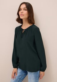 Blusa a maniche lunghe di colore verde scuro con scollatura a chiave e dettaglio a fiocco, realizzata in tessuto morbido; vestibilità rilassata con maniche arricciate.