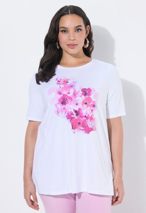 Kvinde med langt mørkt hår iført en hvid kortærmet skjorte med pink blomsterdesign og lyserøde bukser, stående mod en hvid baggrund.