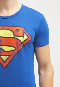 Niebieski bawełniany t-shirt z przetartym logo Supermana w kolorach żółtym i czerwonym, okrągły dekolt i krótkie rękawy. Lekka tekstura z dopasowanym krojem.