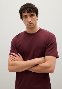 Camiseta de algodón burdeos con mangas cortas, cuello redondo y un bolsillo en el pecho. La tela parece suave y tiene un corte clásico.