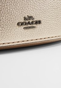 Metaliczna złota torebka z fakturowanej skóry z tłoczonym logo "COACH NEW YORK" i tonującym przeszyciem wzdłuż krawędzi.