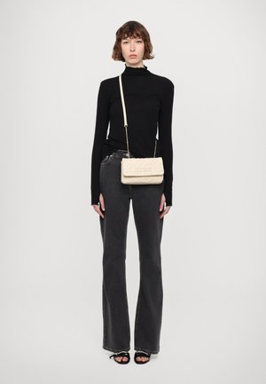 Borsa a tracolla beige trapuntata con una tracolla dorata. Il modello indossa un top nero a maniche lunghe e jeans svasati scuri. Sfondo semplice.