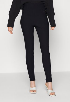 Femme portant un pantalon noir ajusté et des sandales blanches à talons ouverts, se tenant devant un fond uni.