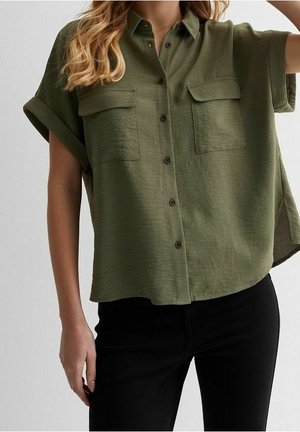 Kobieta ubrana w luźną oliwkowozieloną koszulę na krótki rękaw z dwoma kieszeniami na piersi oraz czarne spodnie na jednolitym tle.