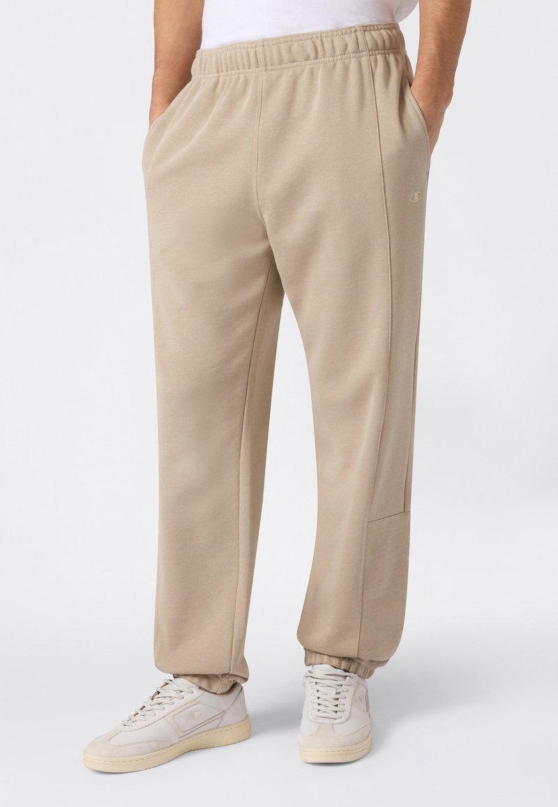 Pantalon de survêtement beige en tissu doux avec une taille élastique, des poches latérales et des poignets côtelés, associé à des baskets de couleur claire.