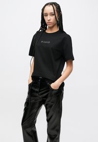 Μαύρο βαμβακερό t-shirt με λογότυπο "PINKO", σε συνδυασμό με μαύρο, γυαλιστερό, ψηλόμεσο παντελόνι. Το μοντέλο έχει μακριά πλεξούδες και τα χέρια στις τσέπες.