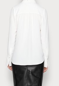Blusa bianca a maniche lunghe con un yoke posteriore plissettato, dettagli sui polsini con bottoni e tessuto leggero e liscio. Abbinata a una gonna di pelle nera.