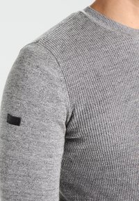 Maglione grigio lavorato a maglia con un motivo testurizzato, collo a giro, e una piccola etichetta nera sulla spalla sinistra, dal design aderente.