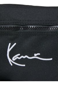 Zwarte stoffen tas met een sluiting met bovenrits en een geborduurd wit handtekening. Materiaal is textuurachtig met een duurzame afwerking.