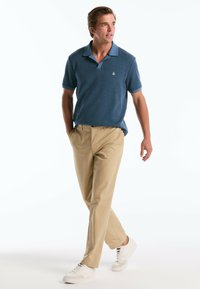 Polo bleu texturé avec un col bleu clair, un pantalon beige et des baskets blanches, arborant un petit logo sur la poitrine.