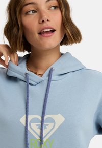 Sudadera azul claro hecha de tela suave, que presenta un cierre con cordón en morado, con un logo de corazón y "ROXY" impreso en verde.