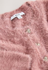 Pūkuotas rausvas kardiganas i�š tekstūruotos, minkštos megztos medžiagos. Papuoštas dekoratyviniais skaidriais akmenų mygtukais ir balta prekės ženklo etikete.