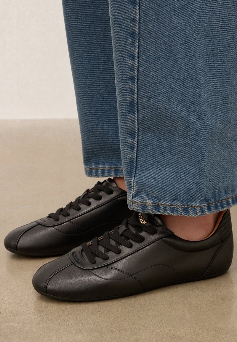 Baskets basses en cuir noir avec des lacets noirs portées avec un jean denim bleu clair à jambes larges sur une surface beige.