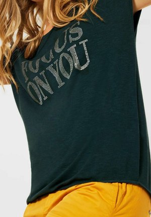 Femme aux cheveux blonds portant une chemise vert foncé avec un texte argenté "FOCUS ON YOU", associée à un pantalon jaune vif.