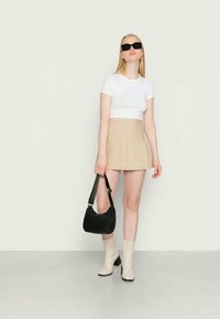 Mulher a usar óculos de sol pretos, t-shirt curta branca, saia quadriculada bege e botas de tornozelo brancas, segurando uma mala preta contra um fundo simples.