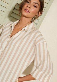 Camicia a righe beige e bianche, con bottoni e colletto, vestibilità rilassata e maniche a metà, realizzata in un tessuto leggero. Orecchini d'oro visibili.