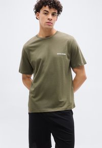 Νέος άνδρας με σγουρά μαλλιά, που φορεί μπλούζα Jack & Jones σε ελιά πράσινο χρώμα και μαύρο σορτς, στέκεται μπροστά σε απλό ανοιχτόφωτο φόντο.