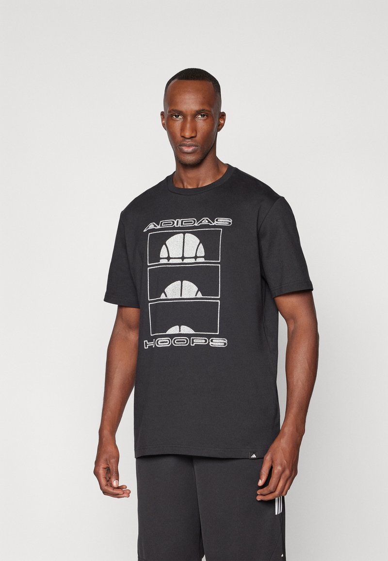 Crna pamučna majica s grafikom koja prikazuje tri dizajna košarkaške lopte i tekst "ADIDAS HOOPS" u bijeloj boji. Kratki rukavi i okrugli izrez.
