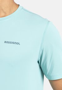 Světle modré sportovní tričko vyrobené z hladkého materiálu. Má kulatý výstřih a krátké rukávy, s potiskem loga "ROSSIGNOL" na přední straně.