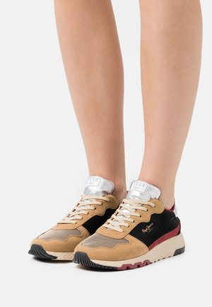 Pieds portant des baskets à lacets beige, noires et bordeaux avec des semelles couleur crème et des languettes rembourrées argentées.