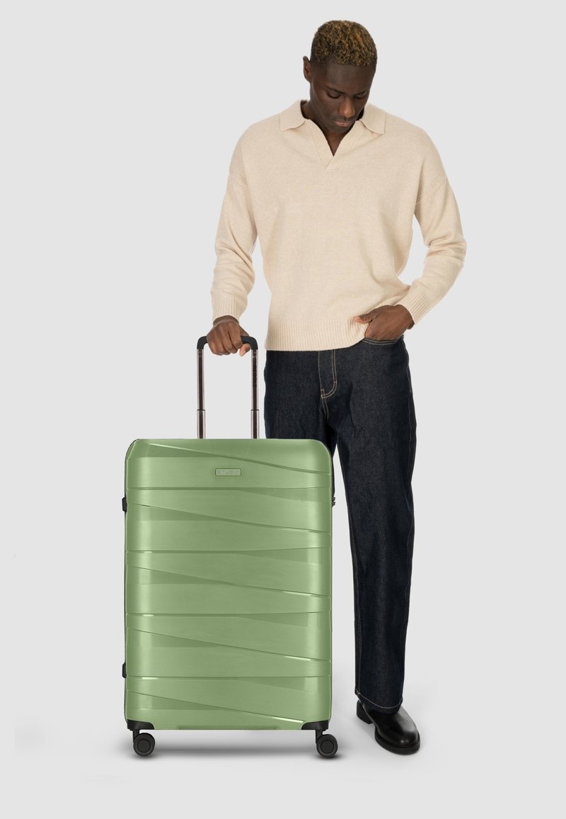 Grüner Hartschalenkoffer mit einem strukturierten Muster, 4 Rädern und einem ausziehbaren Griff, positioniert neben einer Person, die einen cremefarbenen Pullover und Jeans trägt.