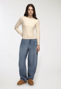 Młoda kobieta z długimi, ciemnymi włosami, w kremowym swetrze w warkocz, luźnych niebieskich dżinsach i brązowych butach wsuwanych, na jednolitym tle.