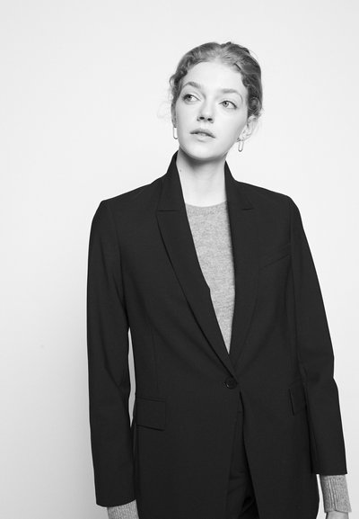 Blazer preto ajustado com fecho de um único botão e bolsos, usado sobre um suéter cinza claro, apresentando uma textura suave e uma forma estruturada.