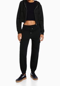 Felpa nera a maniche lunghe con zip, abbinata a pantaloni cargo neri con polsini elasticizzati e tasche, completata da sneakers nere con suole in gomma.