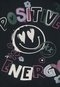 Schwarzer Stoff mit bunten Buchstaben, die "POSITIVE ENERGIE" um ein weißes Smiley-Gesicht mit herzförmigen Augen und dekorativen Blumen anordnen.