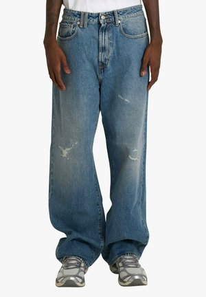 Jeans in denim blu con vestibilità ampia, caratterizzati da toppe consumate, vita alta e design classico a cinque tasche. Indossati con scarpe sportive grigie.