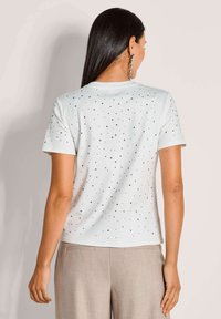 Weißes T-Shirt mit kurzen Ärmeln, das mit verstreuten silbernen Nieten verziert ist. Weicher Stoff mit klassischer Passform, schlichter Rückseite und Rundhalsausschnitt.