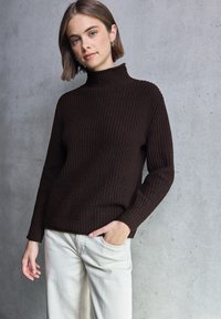 Bruine gebreide coltrui met ribbelstructuur, gecombineerd met lichtgekleurde wijde broek. Model staat tegen een betonnen achtergrond.
