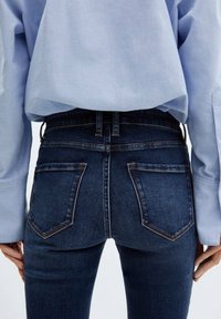 Jeans de tiro alto en azul oscuro con una silueta ajustada, costuras contrastantes y dos bolsillos traseros, combinados con una blusa de manga larga en azul claro.