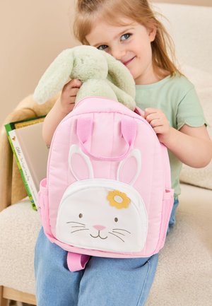 Junges Mädchen hält einen grünen Plüschhasen, während sie einen rosa Rucksack mit weißem Hasengesicht und gelbem Blumendetail packt.