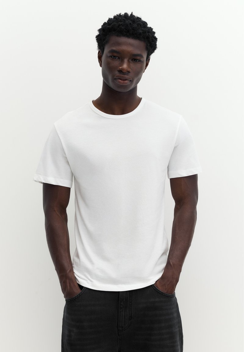 Jeune homme debout sur fond blanc, portant un t-shirt blanc uni et un jean noir, les mains dans les poches.