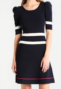 Femme portant une robe bleu marine avec des manches courtes bouffantes, des rayures horizontales blanches sur les manches et la taille, et une fine rayure rouge près de l'ourlet.