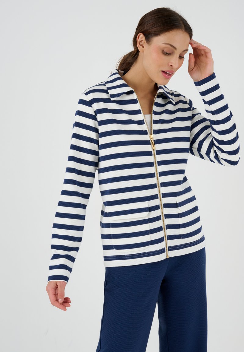 Giacca a zip a righe navy e bianche con colletto, due tasche frontali e dettagli in zip dorati. Materiale misto cotone, vestibilità rilassata.