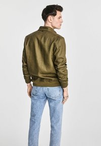 Jasnozielona, materiałowa kurtka z kołnierzem i ściągaczami, zestawiona z jasnoniebieskimi dżinsami. Kurtka ma teksturowaną powierzchnię i luźny krój.